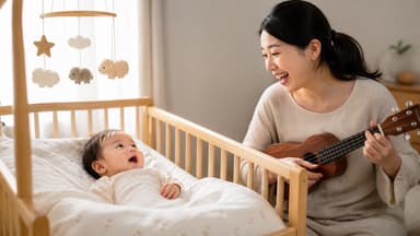 A wide-awake baby lying in a wooden cot under a felt mobile, looking up at her mother who is singing to her with a ukulele