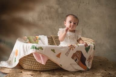 A baby standing at a basket, holding a printed bamboo muslin over the edge
