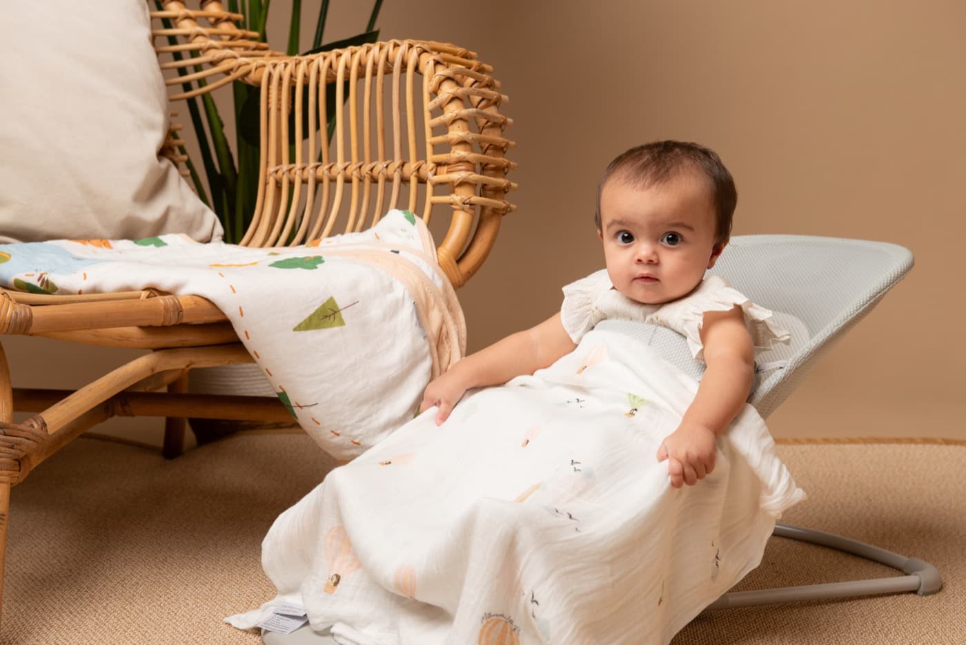 A baby reclining in a bouncer under a bamboo muslin beside a rattan chair