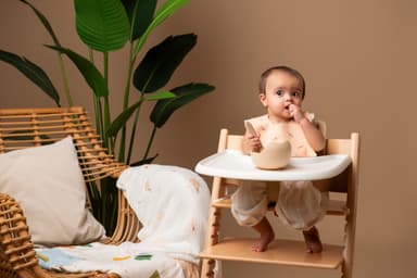 A baby in a wooden high chair feeding herself from a suction bowl