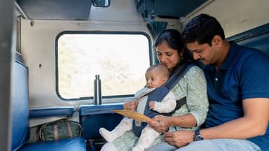 Two parents sitting on a train berth reading a ticket together, their baby upright in a carrier on the mother's chest, a bag stowed beneath the seat