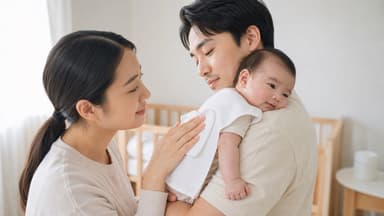A father holding a baby over his shoulder with a white muslin cloth laid across it while the mother pats the baby's back