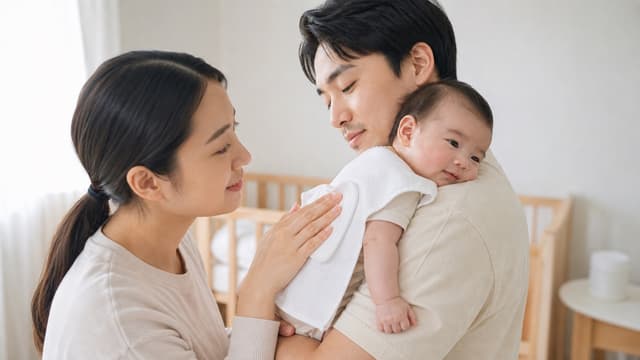 A father holding a baby over his shoulder with a white muslin cloth laid across it while the mother pats the baby's back