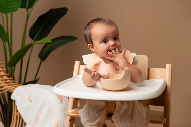 A baby bringing a spoon to her mouth in a high chair, bowl in front of her