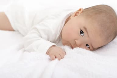 A baby lying on her front on a white blanket with her head turned to one side and both arms free in front of her