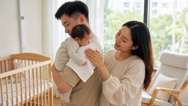 A father holding a baby upright against his shoulder to burp her while the mother rests a hand on her back, in a nursery beside a wooden cot