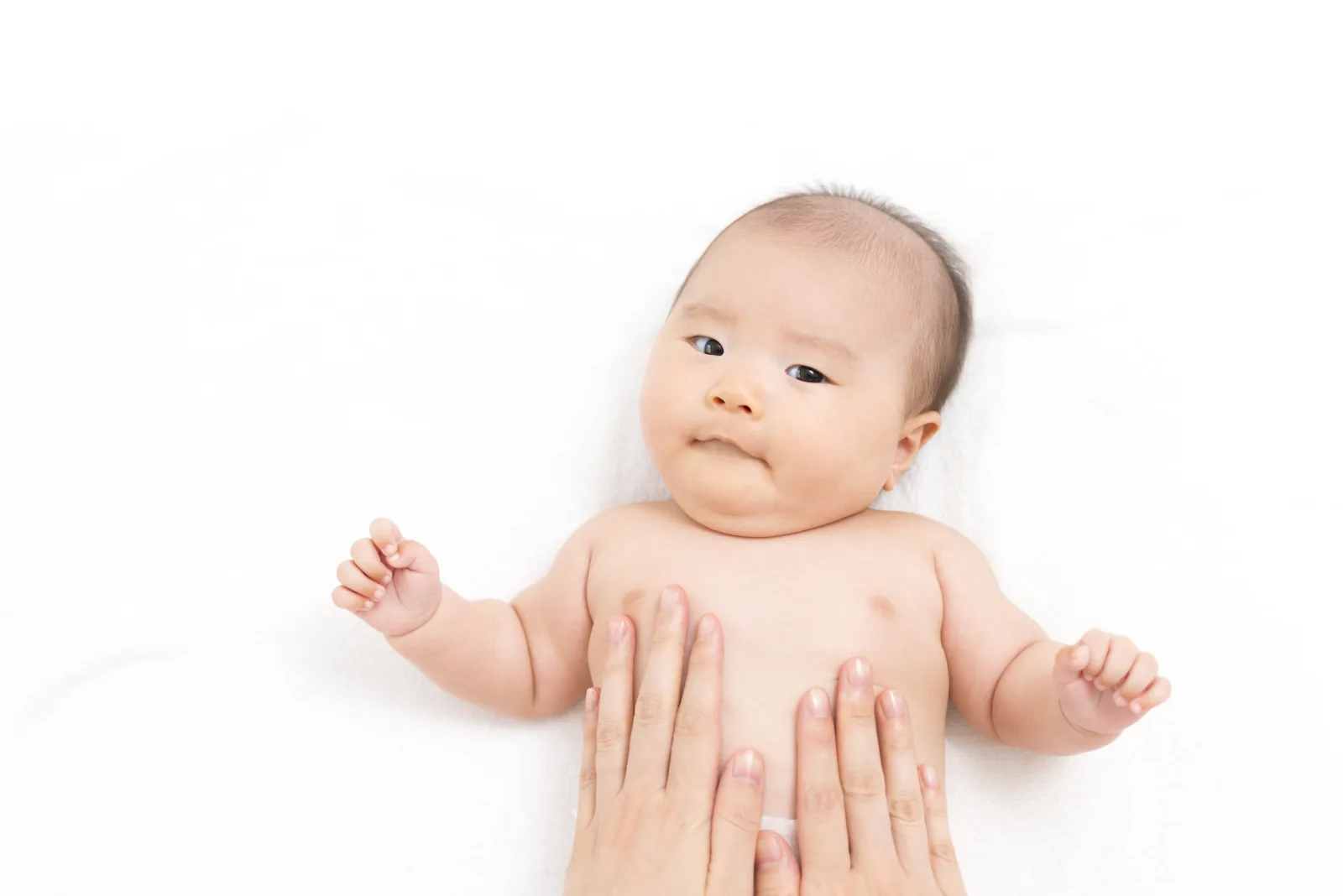 Two adult hands resting flat on a baby's bare tummy to massage it while she lies on her back looking up