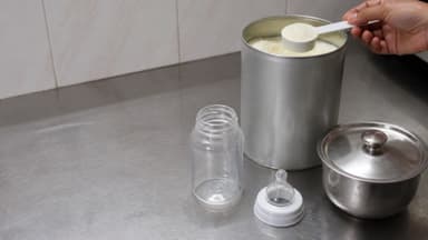 A hand lifting a level scoop of formula powder from a tin on a steel kitchen counter, an empty bottle and teat laid out beside it