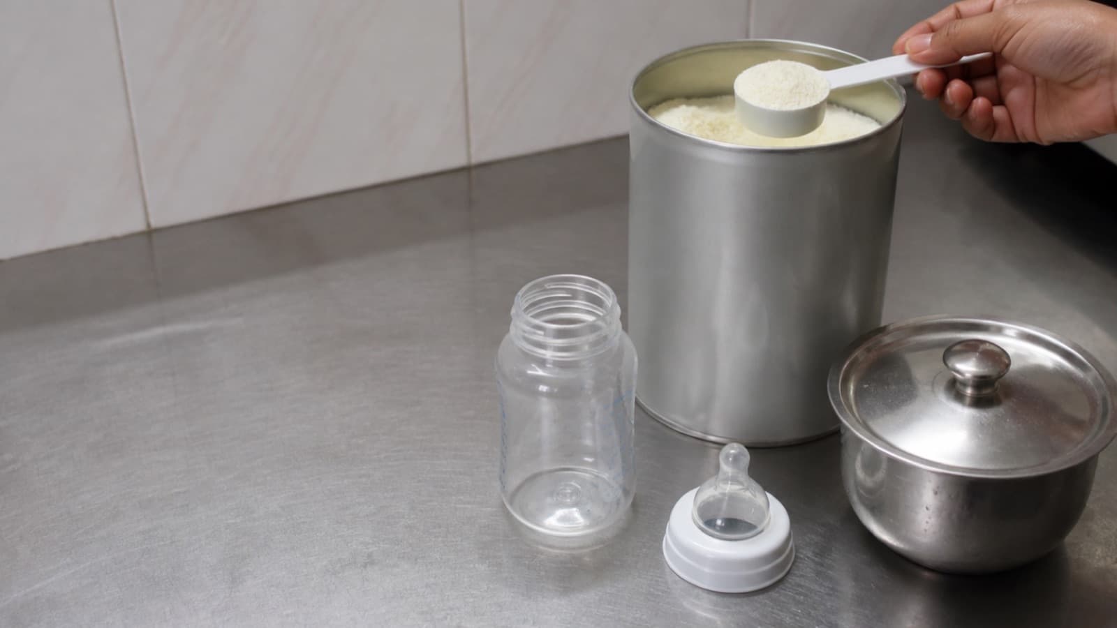 A hand lifting a level scoop of formula powder from a tin on a steel kitchen counter, an empty bottle and teat laid out beside it