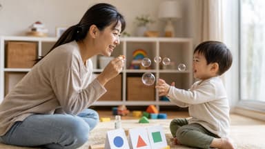 A mother sitting on a playroom rug blowing bubbles towards her toddler, who is laughing and reaching for them, shape cards laid out between them