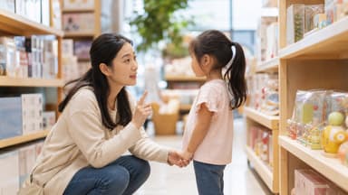 A mother crouched to her daughter's eye level in a shop aisle, holding her hand and explaining something with one finger raised