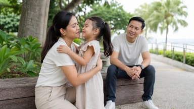 A mother kneeling to her small daughter's eye level to talk with her on a park path, the father sitting on a bench nearby