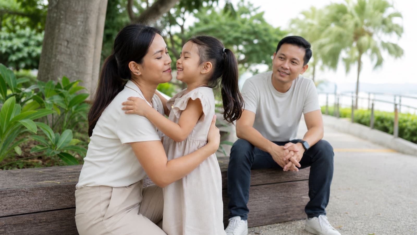 A mother kneeling to her small daughter's eye level to talk with her on a park path, the father sitting on a bench nearby
