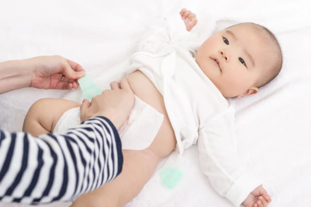 A baby lying on her back on a white towel in an open bodysuit while a parent fastens a fresh nappy