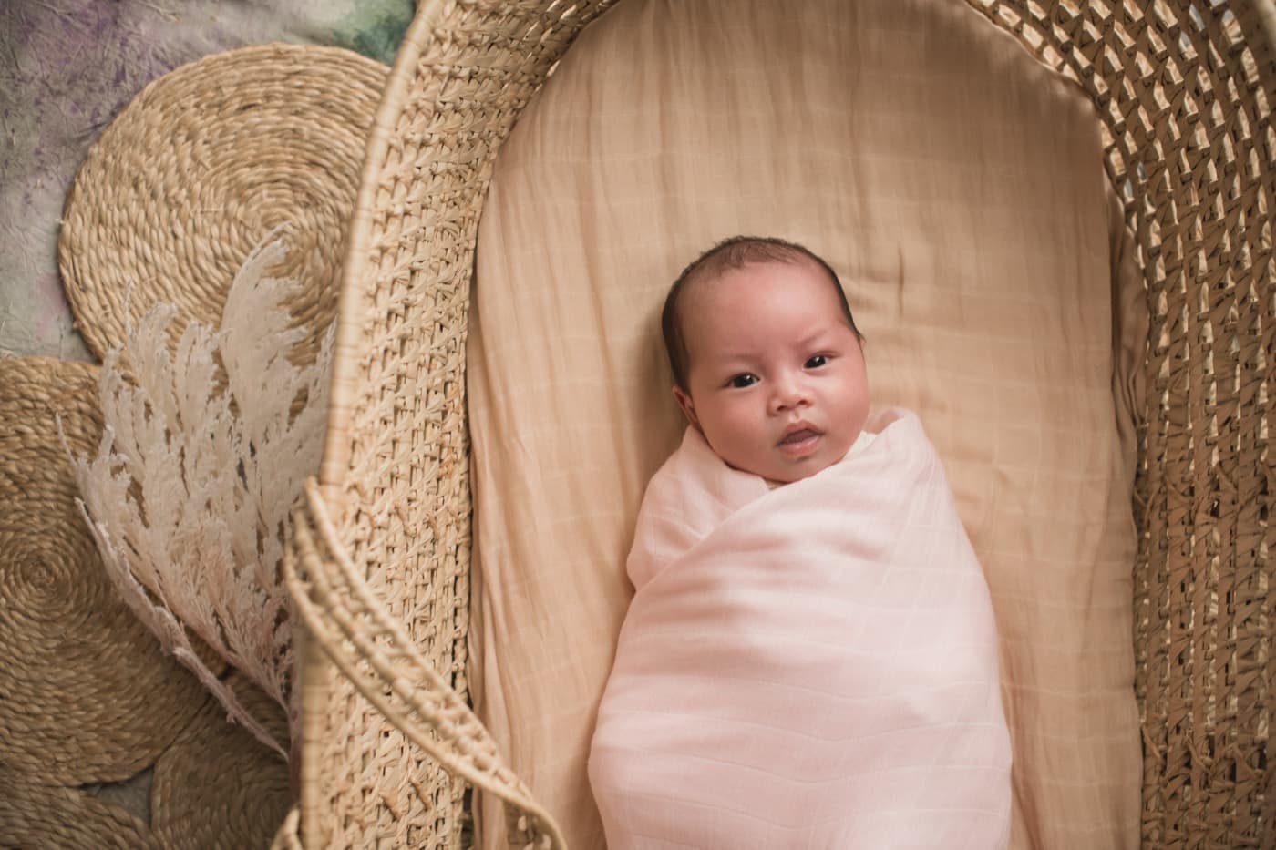 A newborn settled in a woven moses basket in a pale pink bamboo swaddle