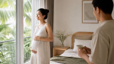 A pregnant woman standing at a bedroom window with both hands resting on her bump, her partner bringing her a cup in the foreground