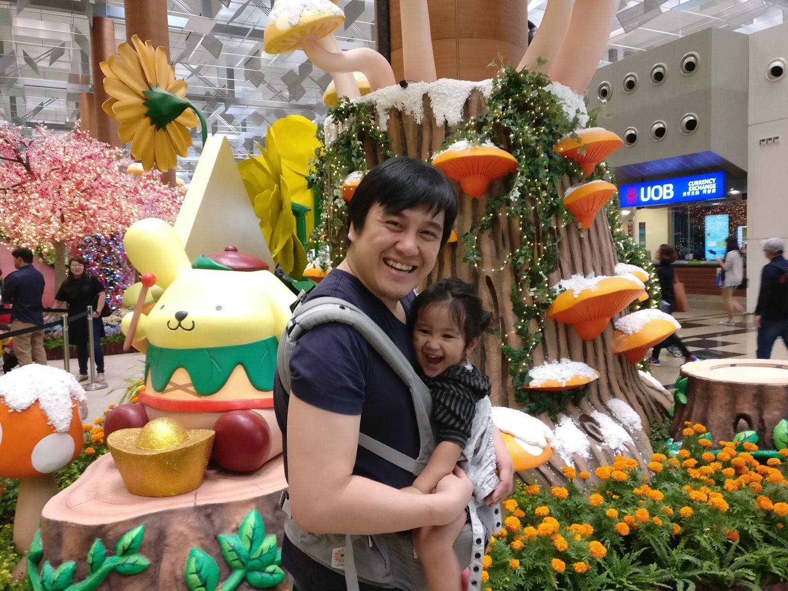Toddler Shan carried chest-to-chest in a structured carrier at Changi Airport, laughing