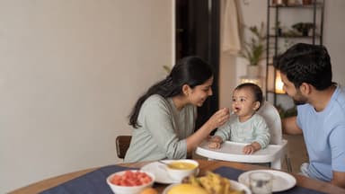 A mother offering a spoonful to a baby sitting upright in a high chair at the family table while the father watches from the other side