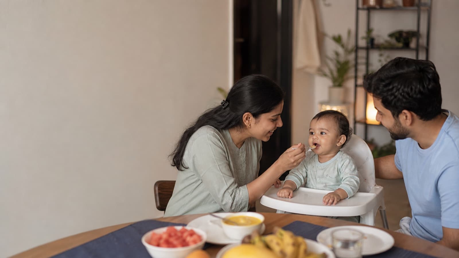 A mother offering a spoonful to a baby sitting upright in a high chair at the family table while the father watches from the other side