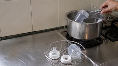 A hand lifting a bottle from a pan of boiling water on a hob, a teat and collar draining in a wire basket beside it