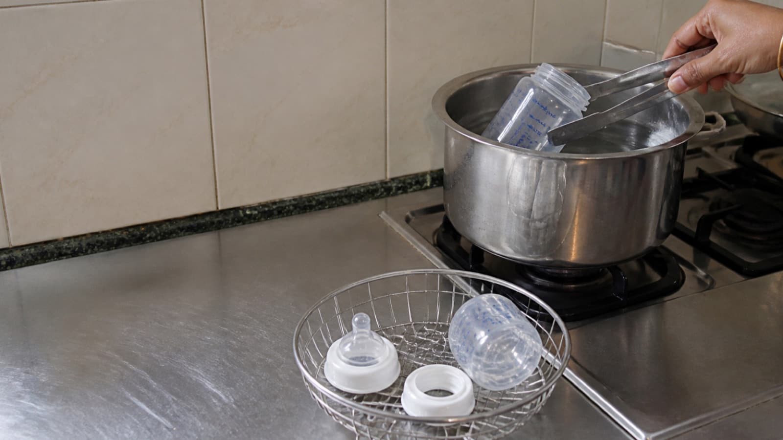 A hand lifting a bottle from a pan of boiling water on a hob, a teat and collar draining in a wire basket beside it