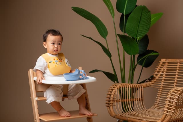 A toddler in a yellow silicone bib at a high chair with a blue bowl and spoon