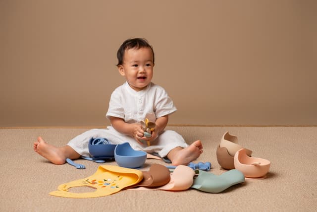 A toddler sitting on the floor surrounded by the Little Rei silicone mealtime range