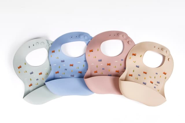 Four Little Rei silicone bibs in sage, blue, rose and sand laid out in a row