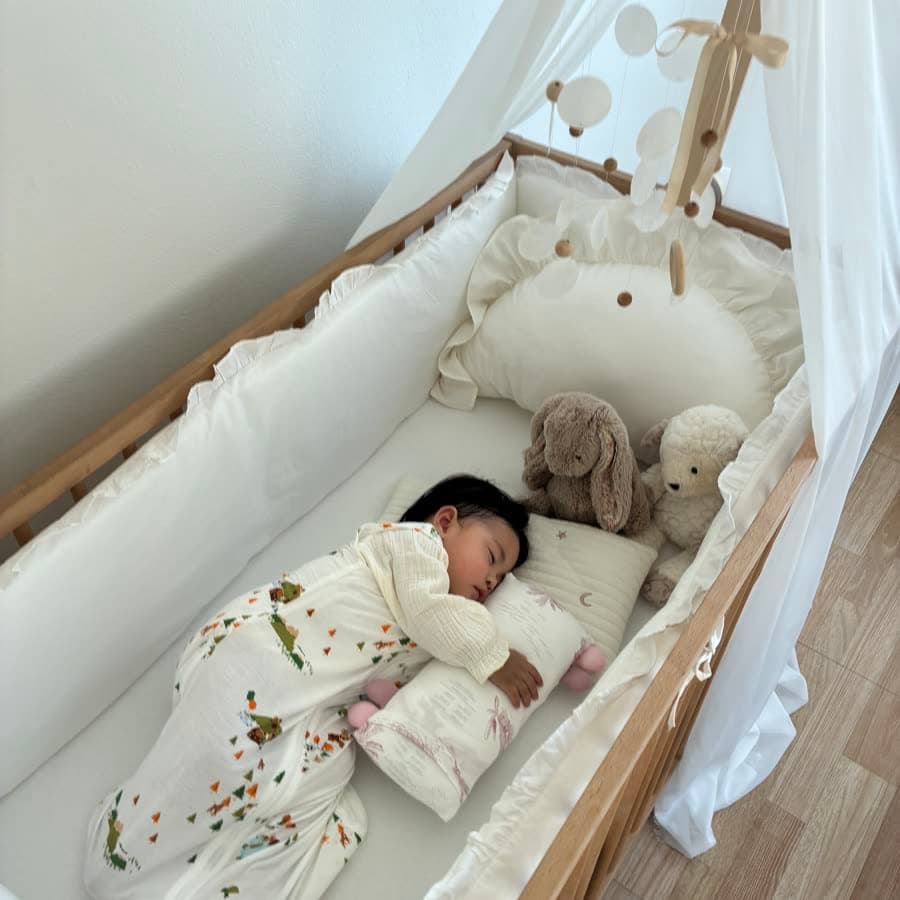 A toddler asleep in a cot alongside a Little Rei pillow and two soft toys