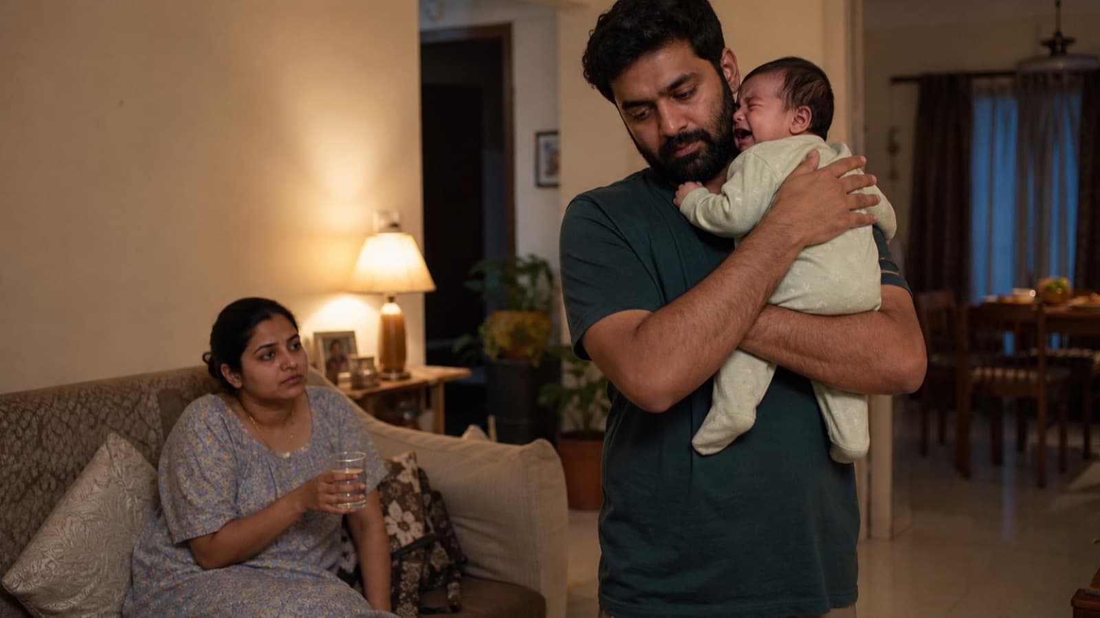 A father standing and holding a crying newborn against his shoulder in a lamplit living room while the mother sits with a glass of water