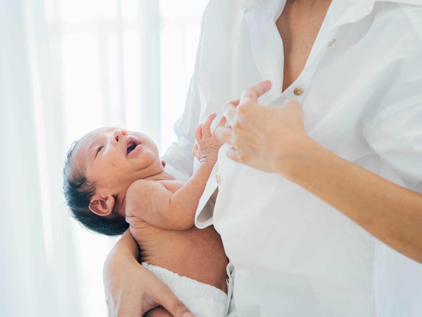 A newborn crying with her mouth open, held against a parent's chest in a bright room, one small hand raised
