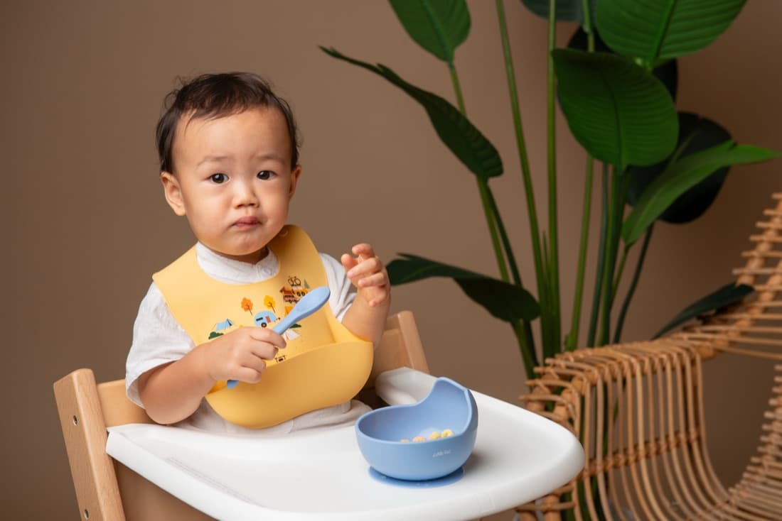 A toddler in a high chair wearing a yellow train bib, holding a spoon over a blue bowl